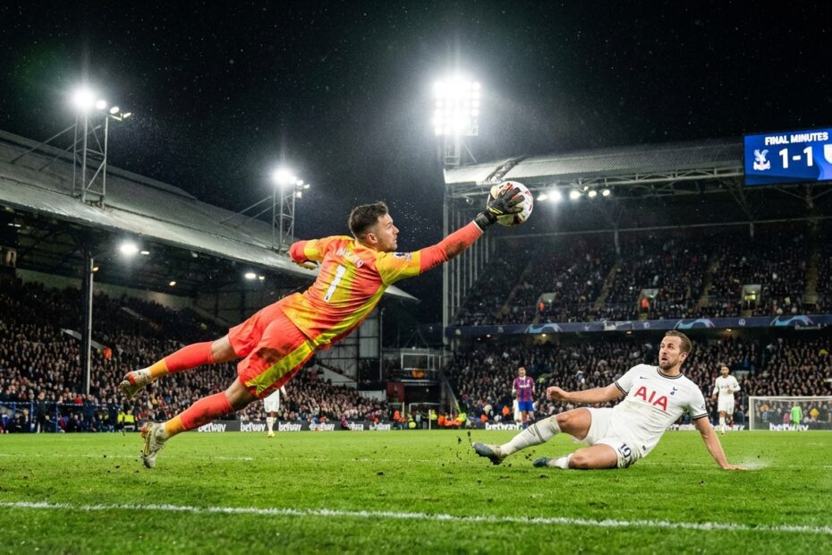 Goleiro salta para fazer defesa milagrosa durante partida de futebol noturna em estádio lotado.